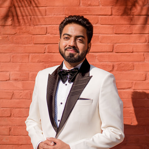Man in a white suit with black lapels standing against a red brick wall with palm leaf shadows.