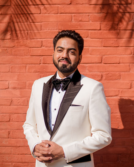 Man in a white suit with black lapels standing against a red brick wall with palm leaf shadows.